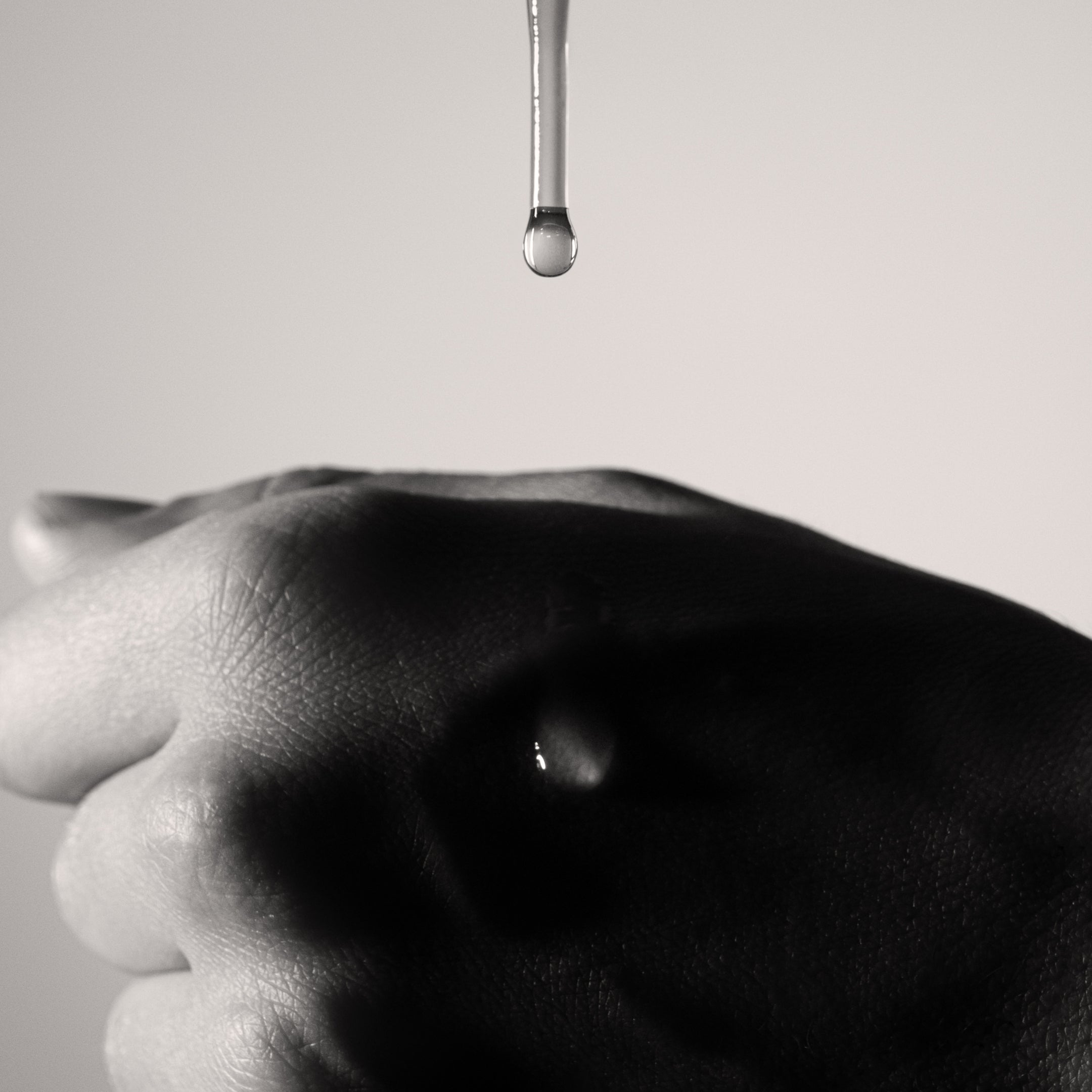 Black and white close-up of a hand with a drop of serum hanging above it against a plain background
